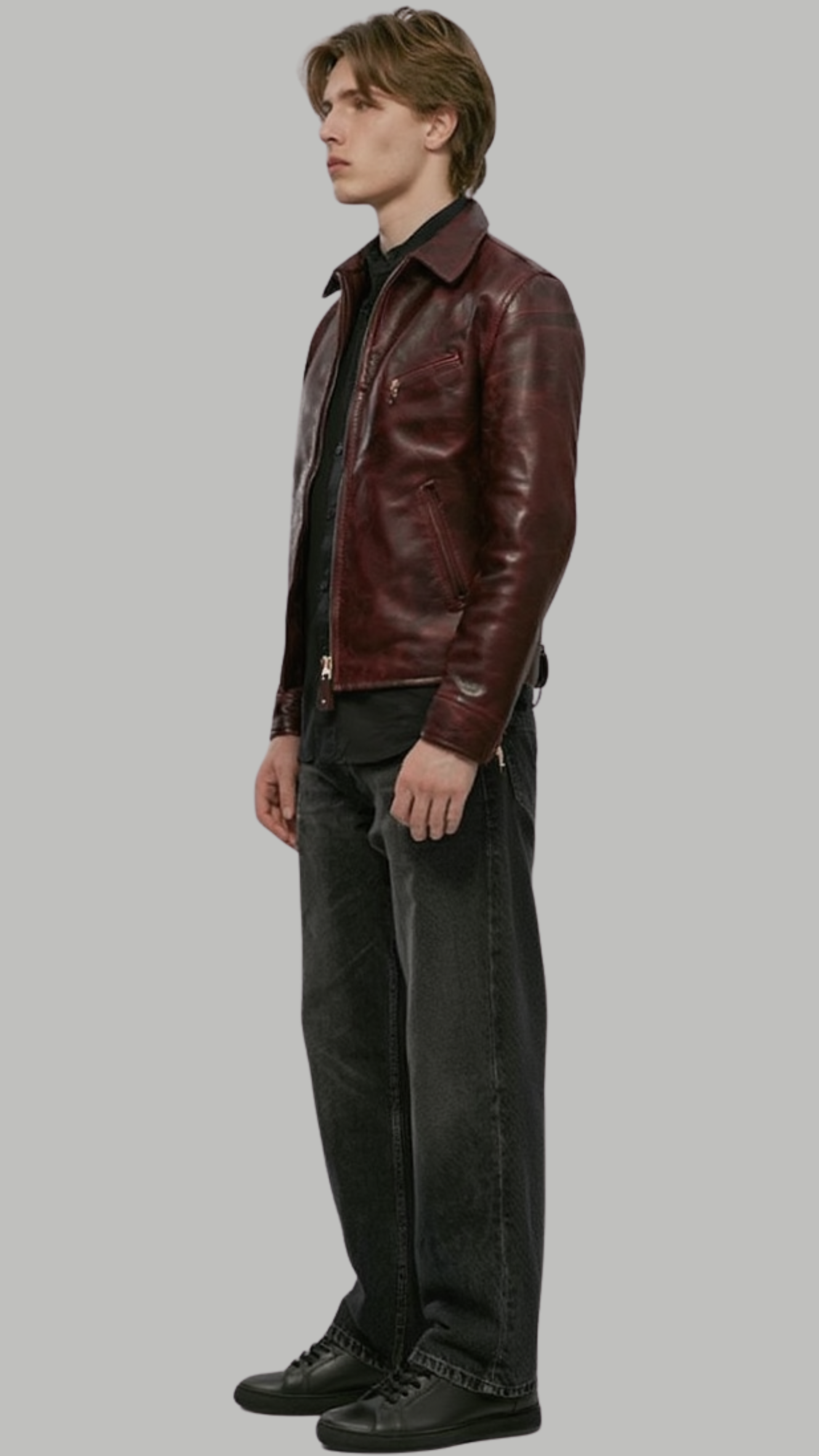 A man wearing a dark burgundy leather jacket, black shirt, black jeans, and black sneakers, standing in profile against a solid gray background.