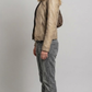 A model wearing a cropped dark brown leather jacket with beige stripes on the chest, paired with dark blue jeans and white sneakers, set against a solid gray background.