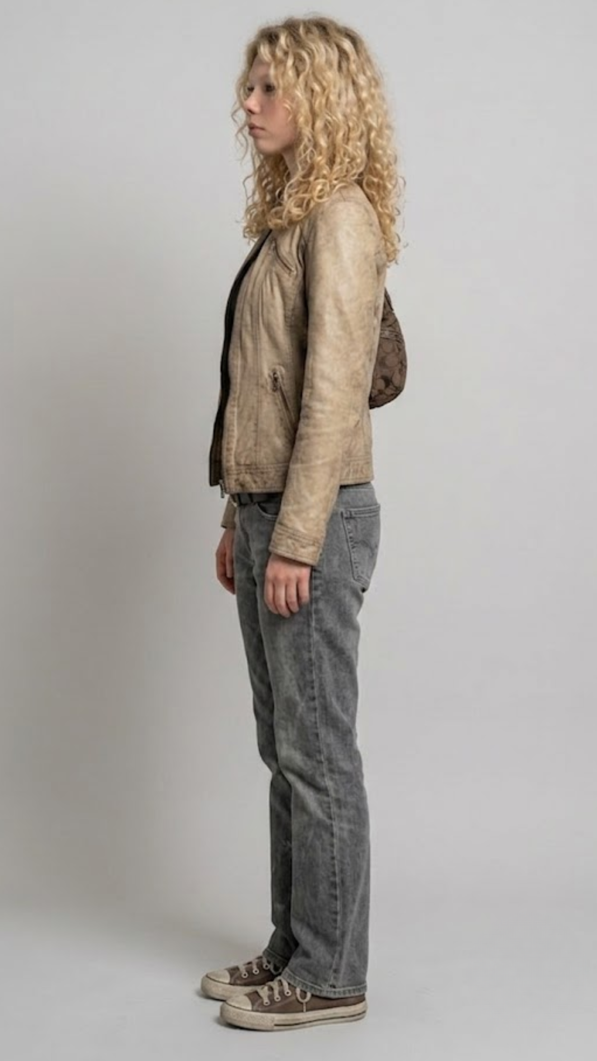 A model wearing a cropped dark brown leather jacket with beige stripes on the chest, paired with dark blue jeans and white sneakers, set against a solid gray background.