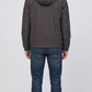 A man viewed from behind wearing a dark gray hooded jacket and blue jeans, standing against a light gray studio background.