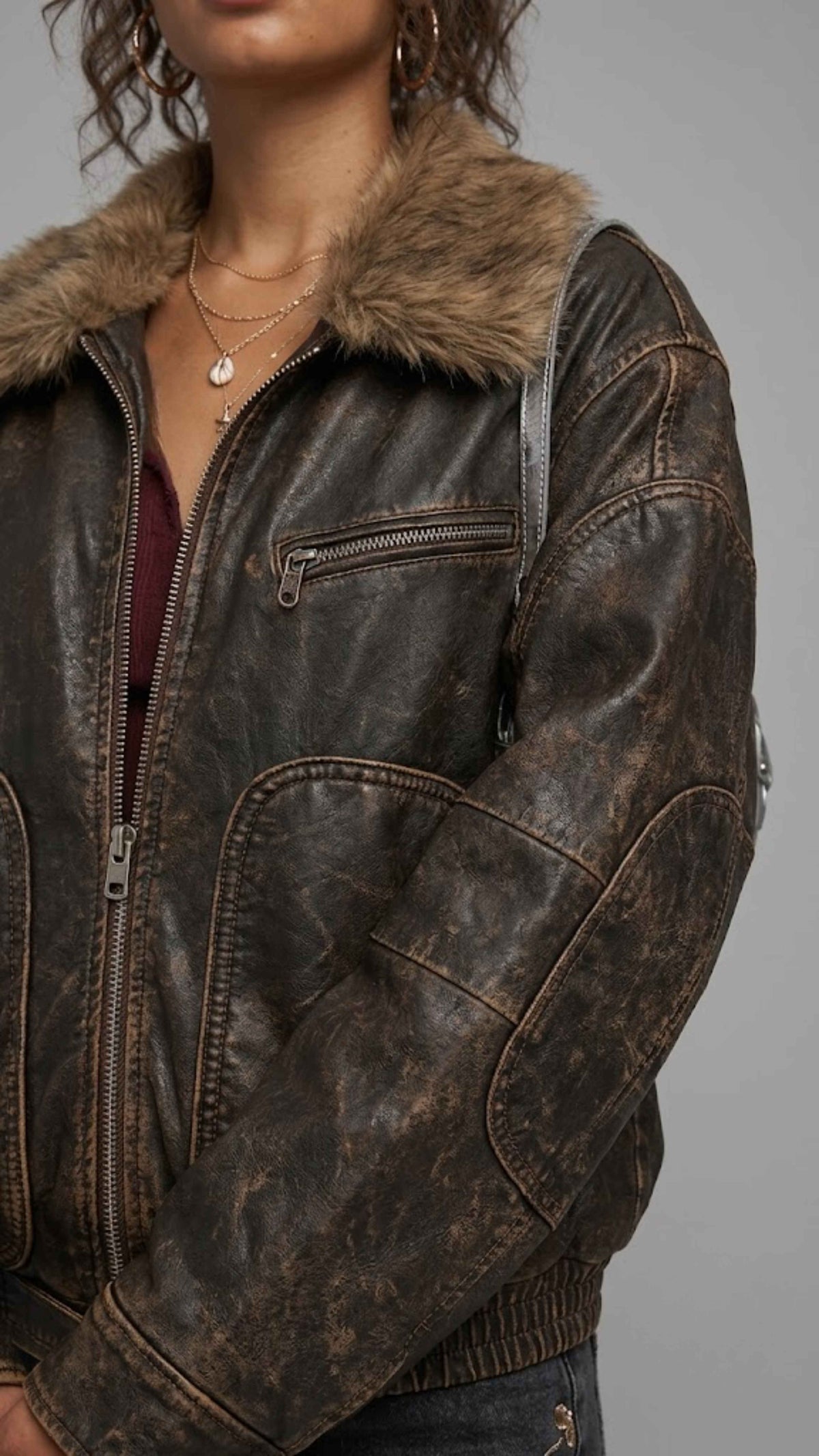 A close-up of a person wearing a distressed dark brown faux-leather jacket with a brown faux-fur collar, shown against a plain gray background.