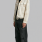 A model wearing a light beige distressed denim jacket over a black t-shirt, paired with black wide-leg jeans and silver sneakers, standing against a solid gray background.