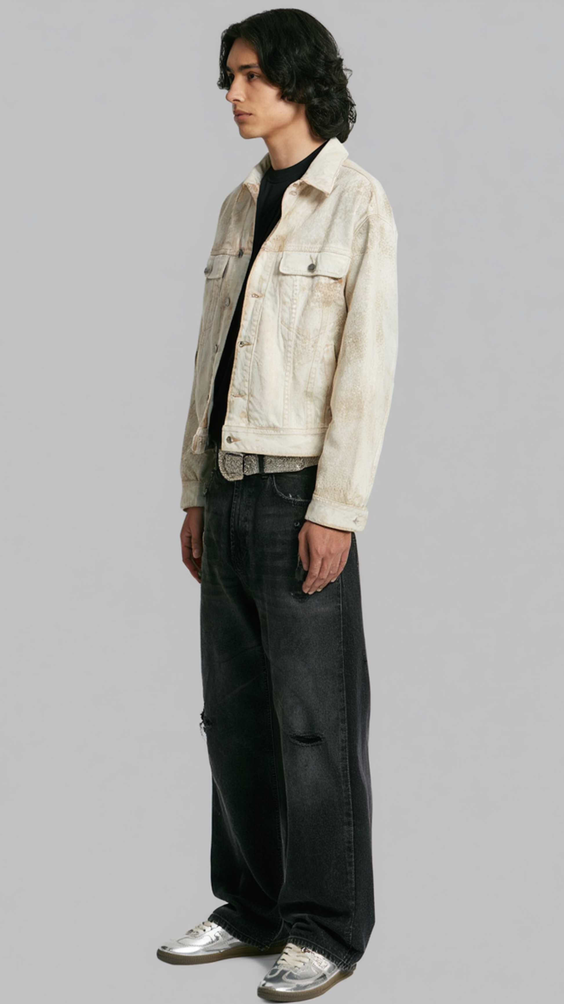 A model wearing a light beige distressed denim jacket over a black t-shirt, paired with black wide-leg jeans and silver sneakers, standing against a solid gray background.