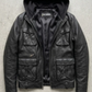 A black leather jacket with an integrated black hood, laid flat on a textured gray concrete background. The jacket features multiple front flap pockets and a zippered chest pocket, with the brand name 'JACKERO' visible on the inner neck label.