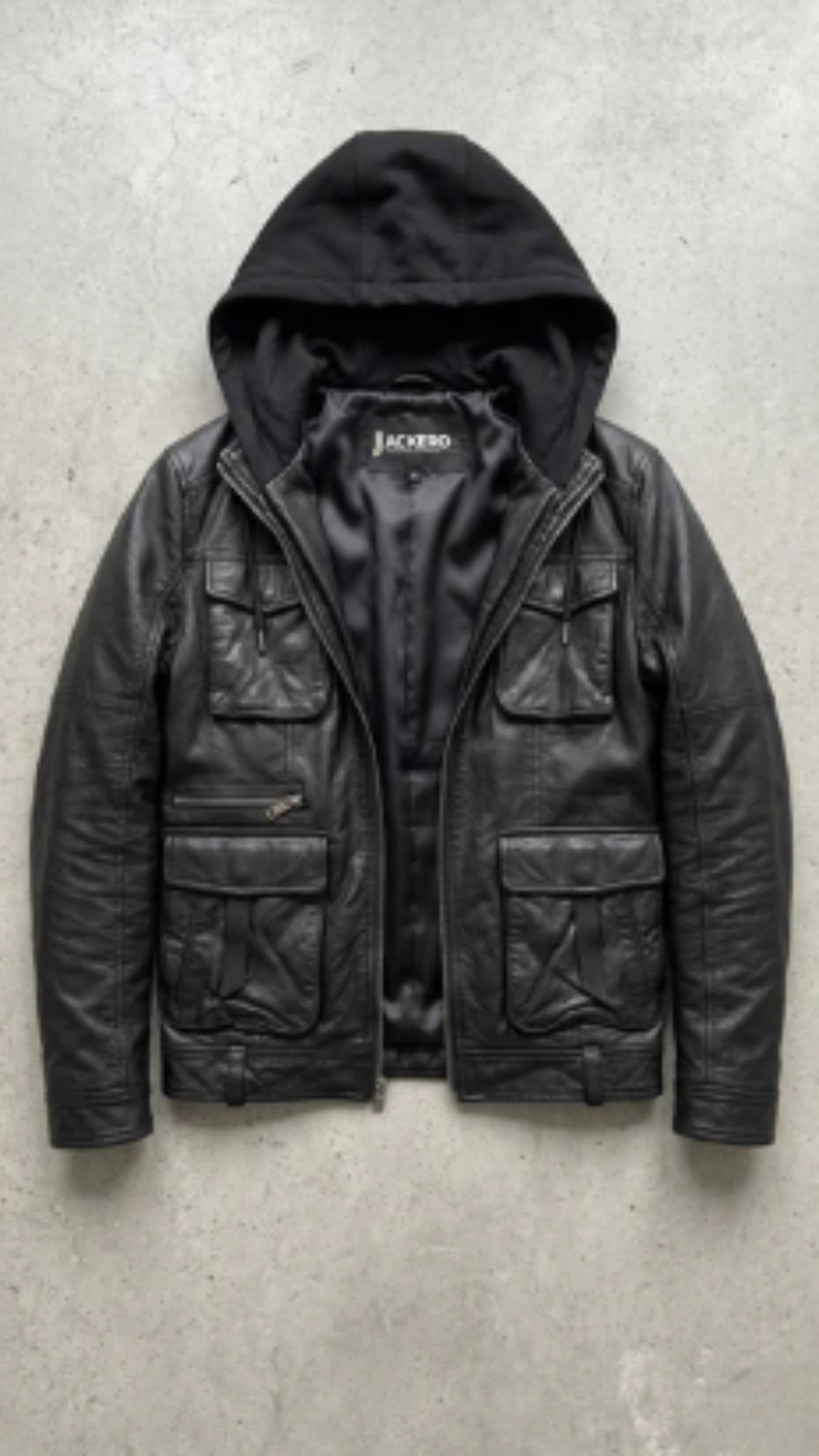 A black leather jacket with an integrated black hood, laid flat on a textured gray concrete background. The jacket features multiple front flap pockets and a zippered chest pocket, with the brand name 'JACKERO' visible on the inner neck label.
