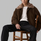 Man sitting on a wooden chair wearing a brown coat with fur collar.