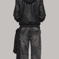 A model shown from the back wearing a JACKERO black leather hooded jacket with a brown fur-trimmed hood, paired with dark gray washed denim jeans and a dark patterned scarf, against a solid gray background.