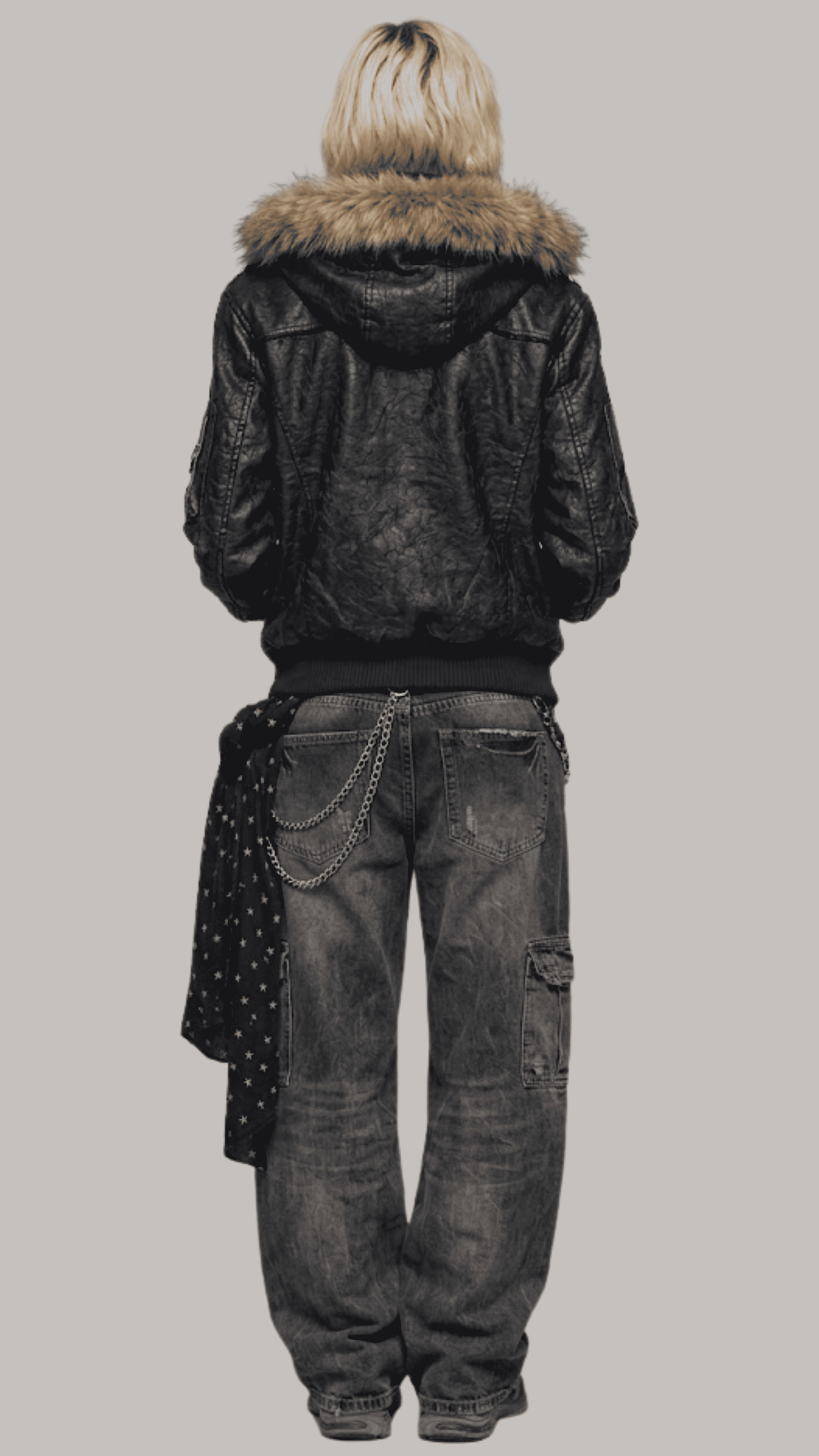 A model shown from the back wearing a JACKERO black leather hooded jacket with a brown fur-trimmed hood, paired with dark gray washed denim jeans and a dark patterned scarf, against a solid gray background.