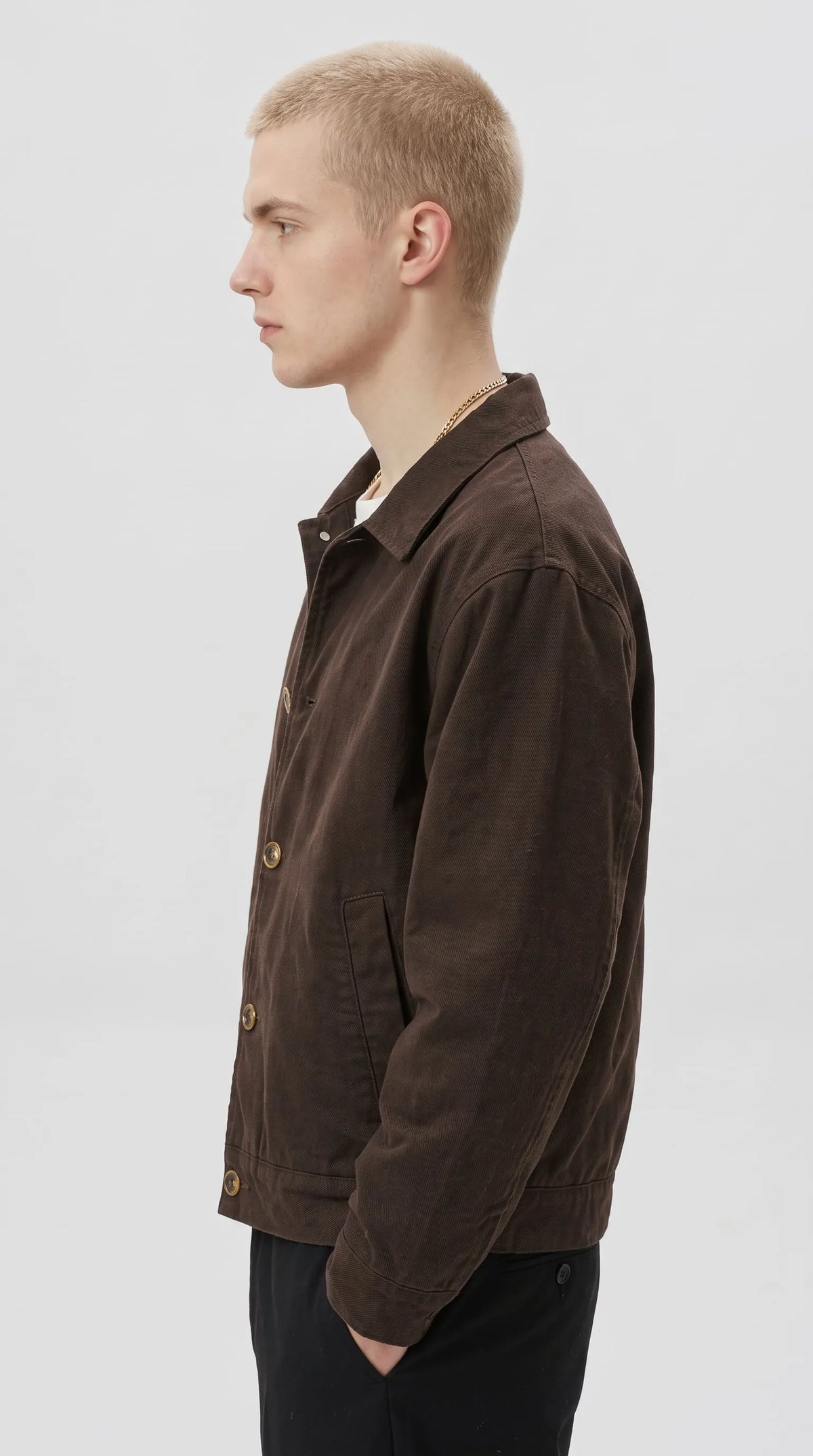 Relaxed dark brown cotton canvas utility jacket with a clean shoulder line, long sleeves, antique-tone button fastening and discreet side-entry pockets, offering a versatile earth-tone streetwear look for everyday outfits.
