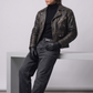 A man wearing a distressed black leather motorcycle jacket, black turtleneck, dark gray jeans, black leather gloves, and black leather shoes, leaning against a minimalist white geometric bench in a studio setting.