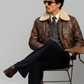 Man sitting on a chair wearing a brown leather jacket with fur lining, dark pants, and sunglasses.
