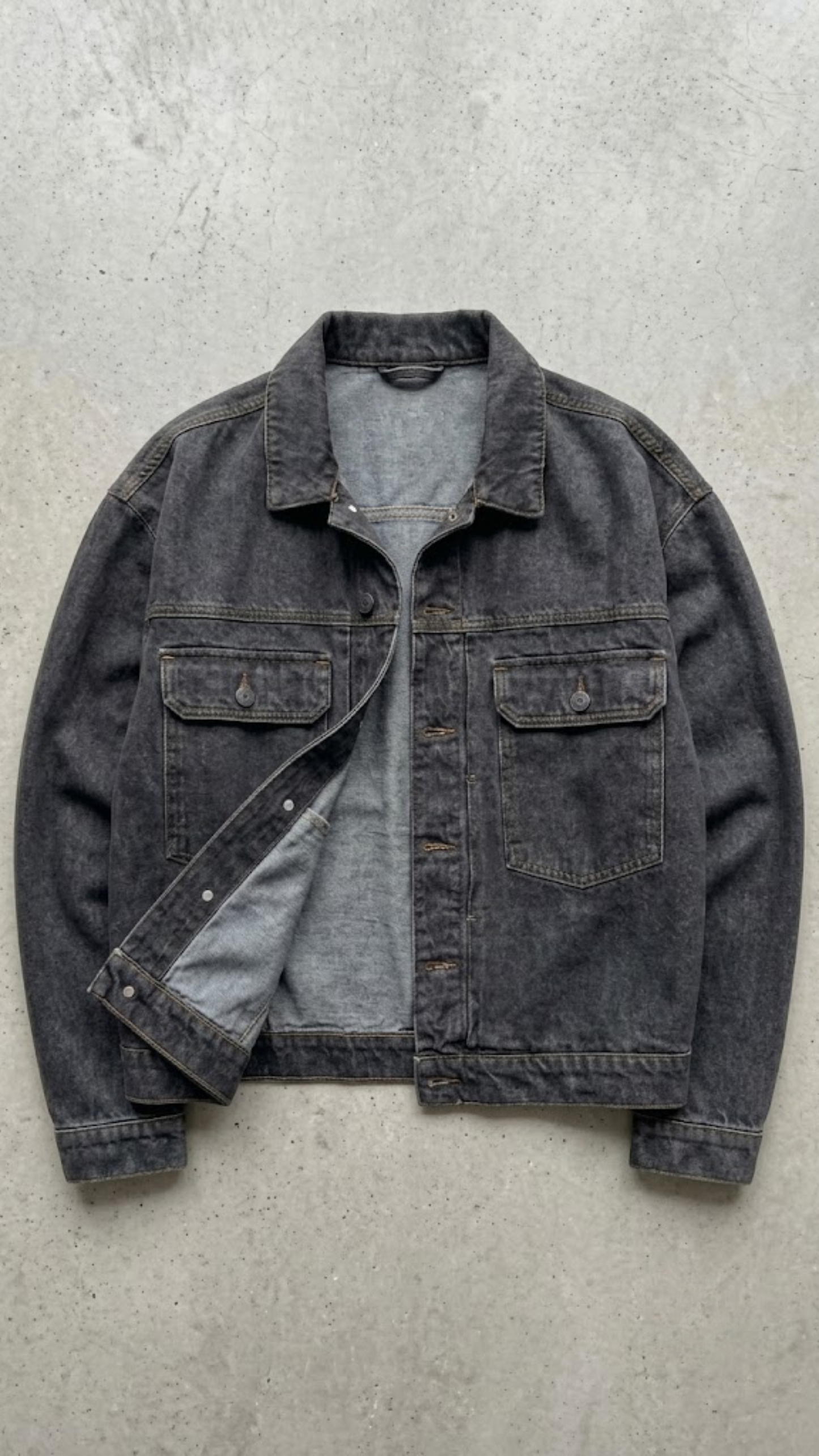 A washed dark charcoal denim jacket, laid flat on a gray concrete surface.