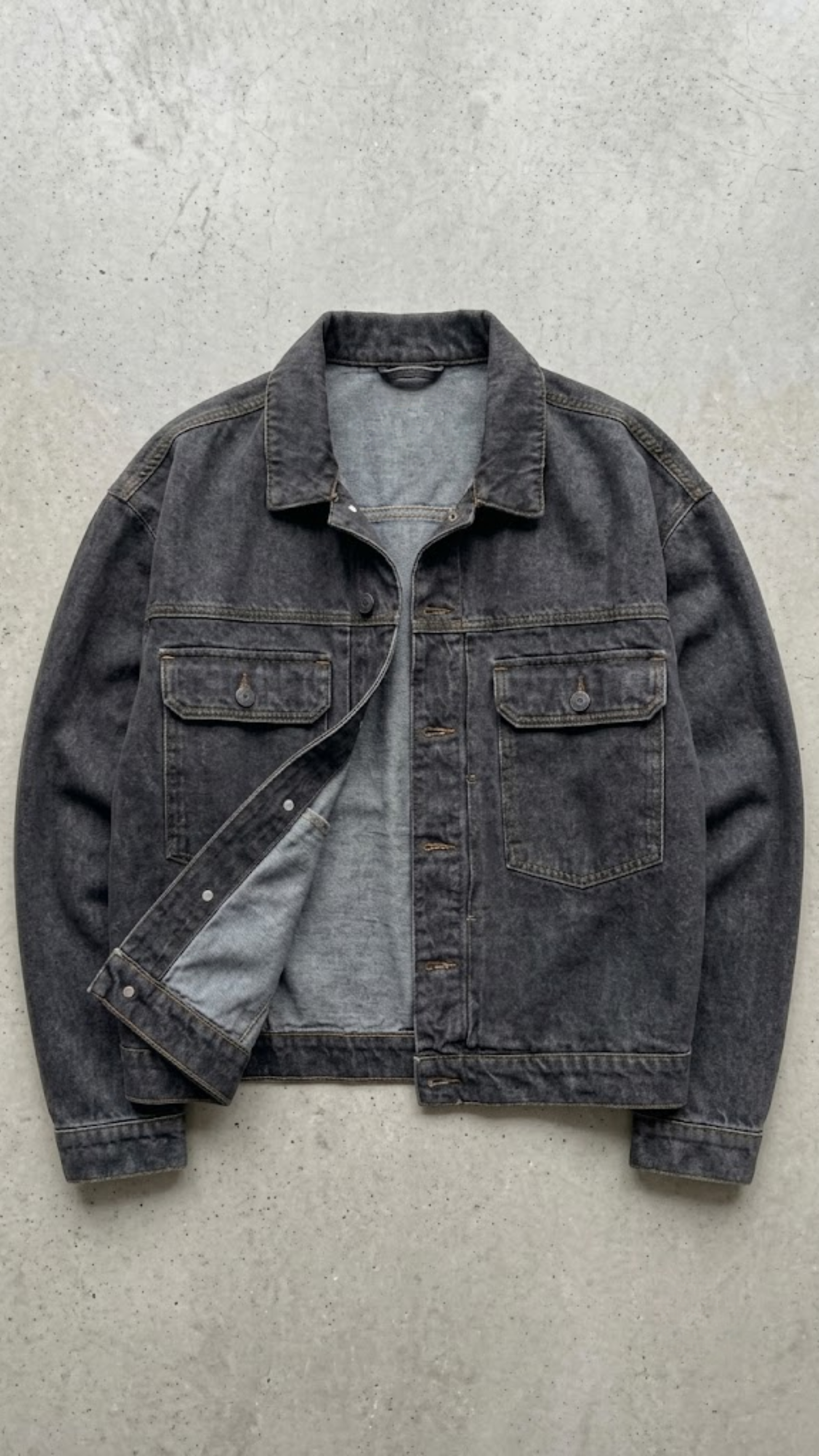 A washed dark charcoal denim jacket, laid flat on a gray concrete surface.