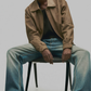 Man sitting on a chair wearing a beige jacket and blue jeans against a plain background
