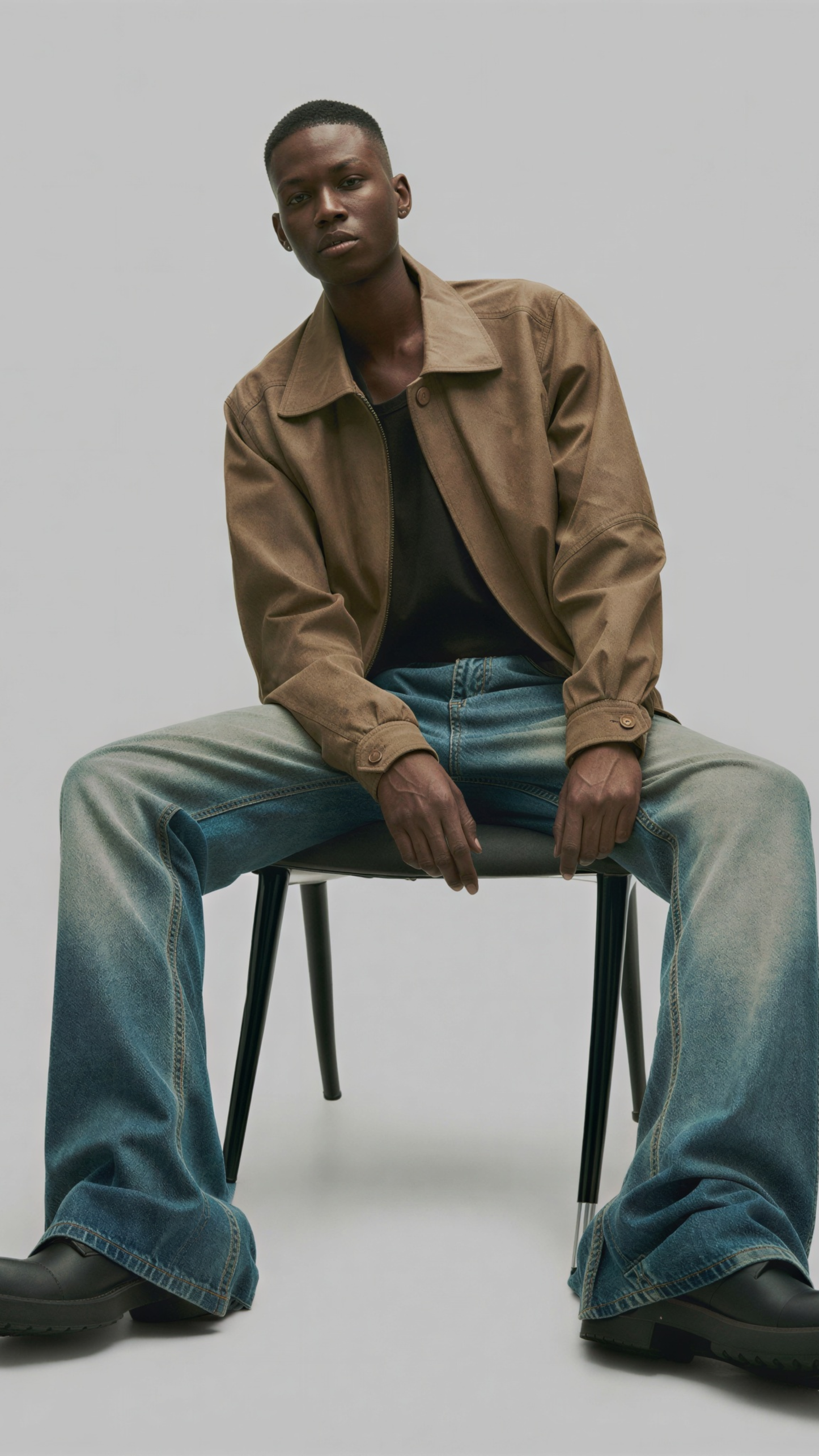 Man sitting on a chair wearing a beige jacket and blue jeans against a plain background