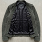 A dark olive green suede jacket with a black satin lining, laid flat on a textured gray concrete surface.