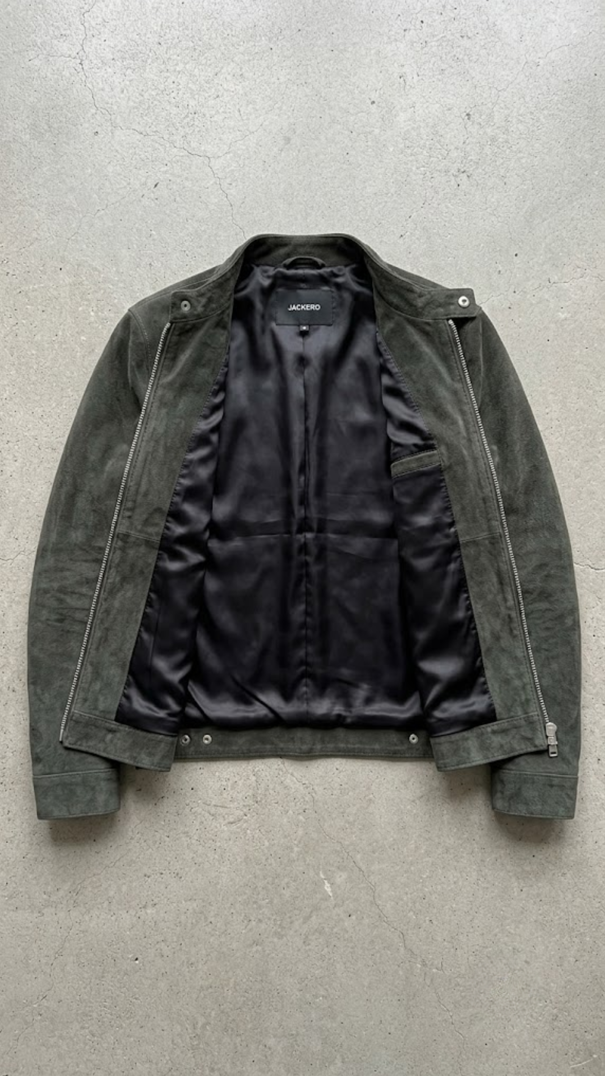 A dark olive green suede jacket with a black satin lining, laid flat on a textured gray concrete surface.