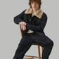 A man with curly brown hair sits on a wooden chair wearing a dark navy jacket with a beige shearling-style collar, matching dark trousers, and black shoes against a solid gray background.