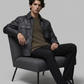 Man sitting on a gray chair wearing a black leather jacket and dark pants against a plain background