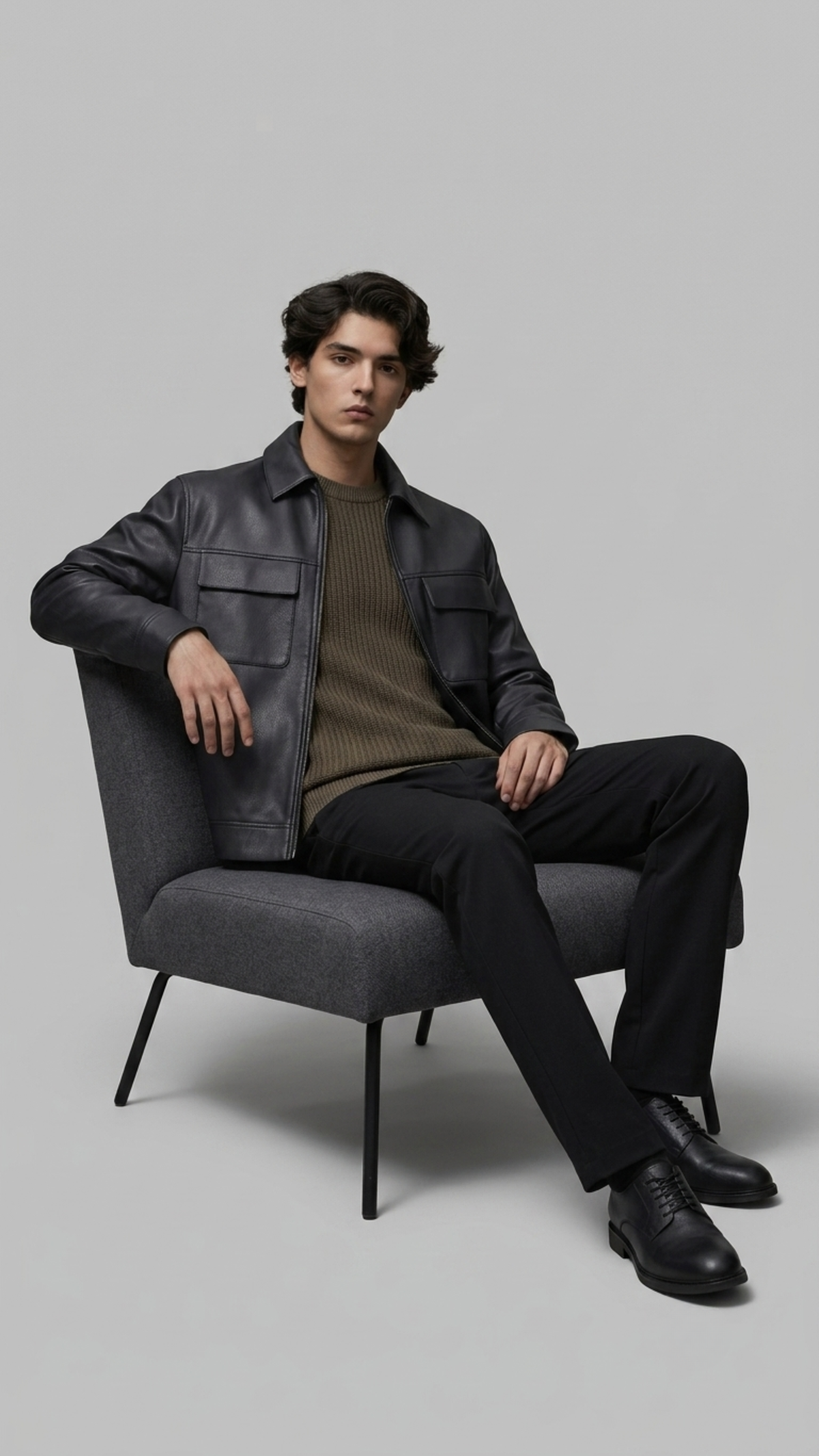 Man sitting on a gray chair wearing a black leather jacket and dark pants against a plain background