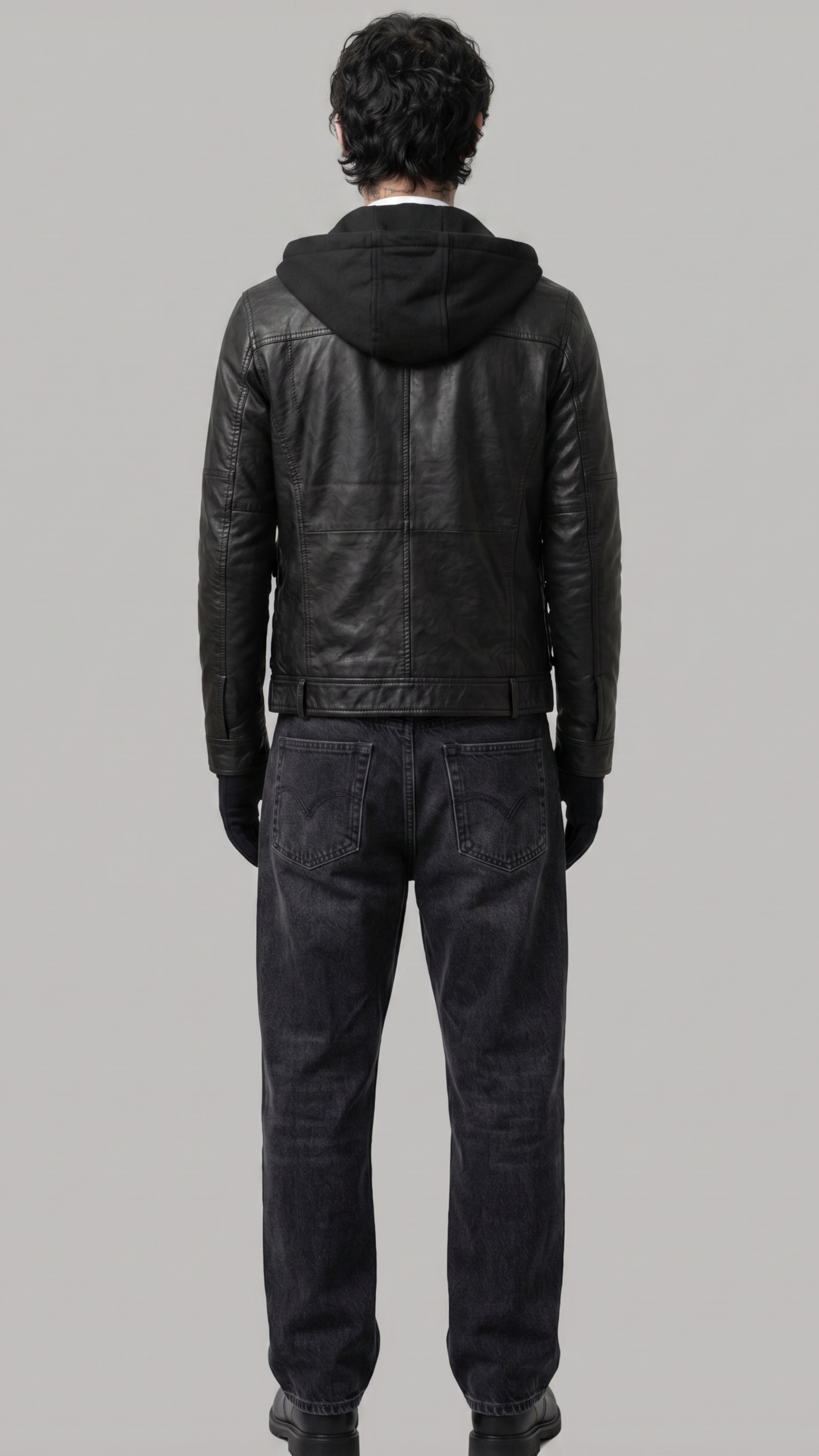 A man viewed from behind wearing a black leather hooded jacket, dark gray jeans, and black gloves against a solid gray studio background.