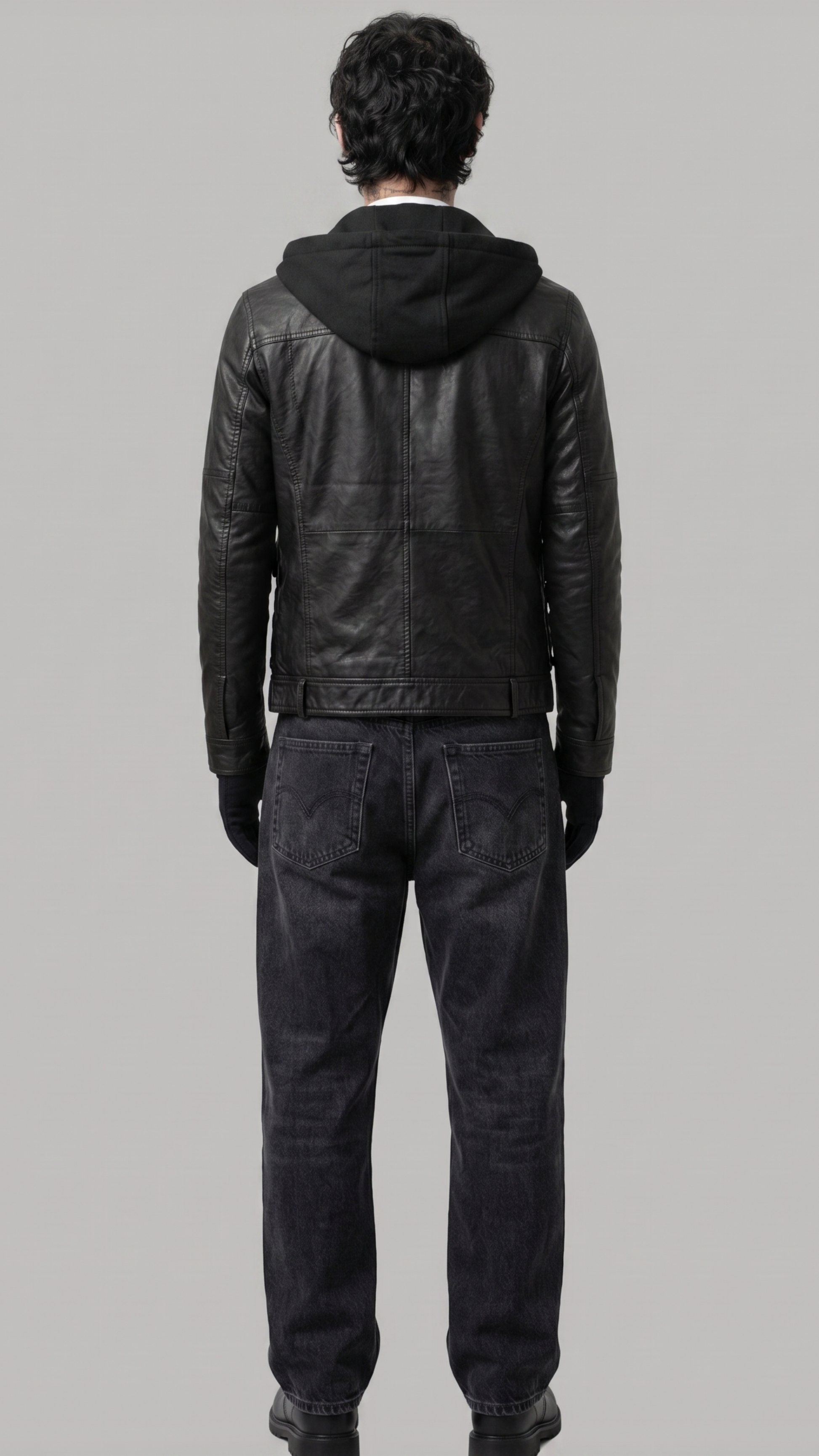 A man viewed from behind wearing a black leather hooded jacket, dark gray jeans, and black gloves against a solid gray studio background.