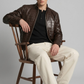 Man sitting on a wooden chair wearing a brown crocodile leather jacket, black shirt, beige pants, and black shoes.
