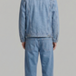 A man viewed from behind wearing a light blue denim jacket and matching light blue denim pants against a plain light gray studio background.