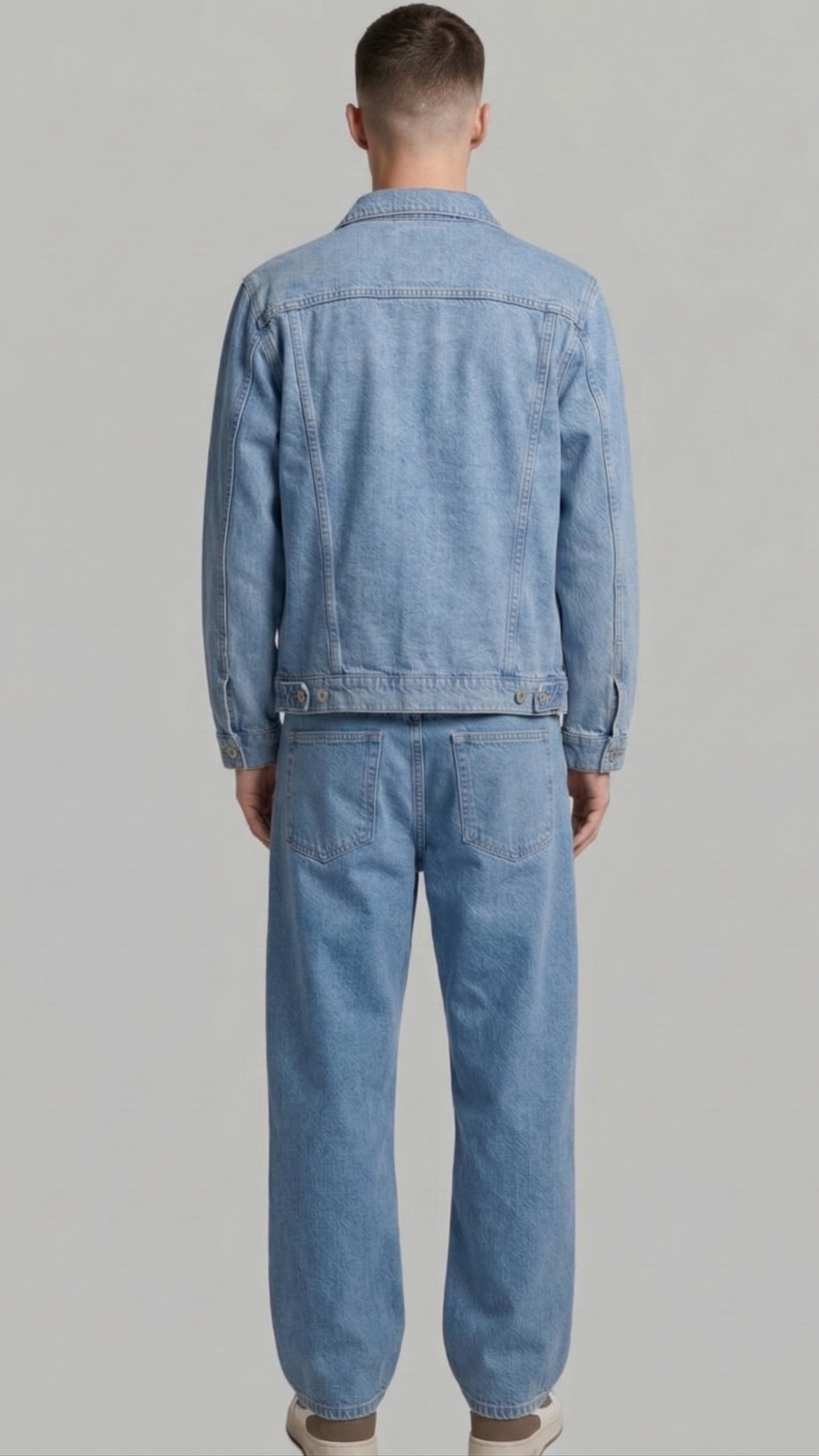 A man viewed from behind wearing a light blue denim jacket and matching light blue denim pants against a plain light gray studio background.