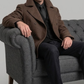 Man in a brown wool pea coat and cap sitting on a gray sofa against a light gray background