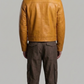 A man viewed from the back wearing a mustard yellow leather jacket and dark brown pants, standing against a solid gray studio background.