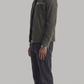 A man in profile wearing a dark olive green suede jacket, black shirt, dark charcoal jeans, and black leather boots, standing against a solid light gray background.