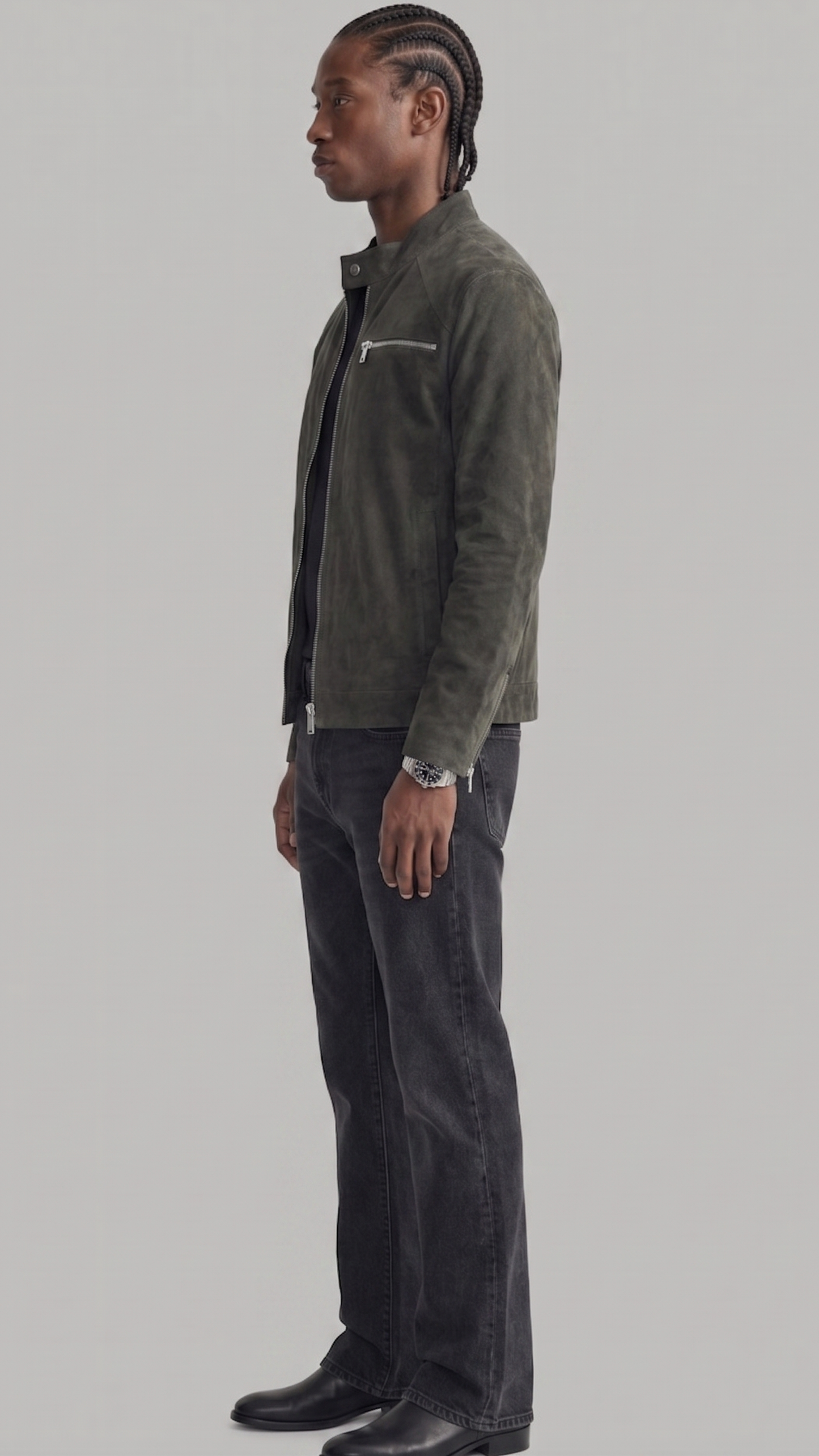 A man in profile wearing a dark olive green suede jacket, black shirt, dark charcoal jeans, and black leather boots, standing against a solid light gray background.
