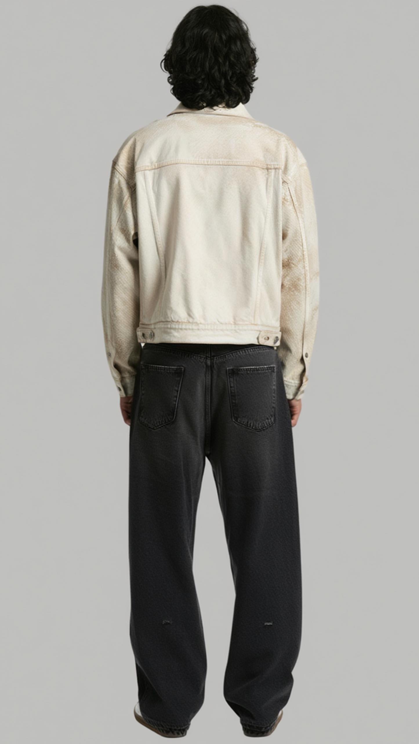 A model shown from the back wearing a cream-colored denim jacket and dark charcoal wide-leg jeans, set against a solid gray studio background.