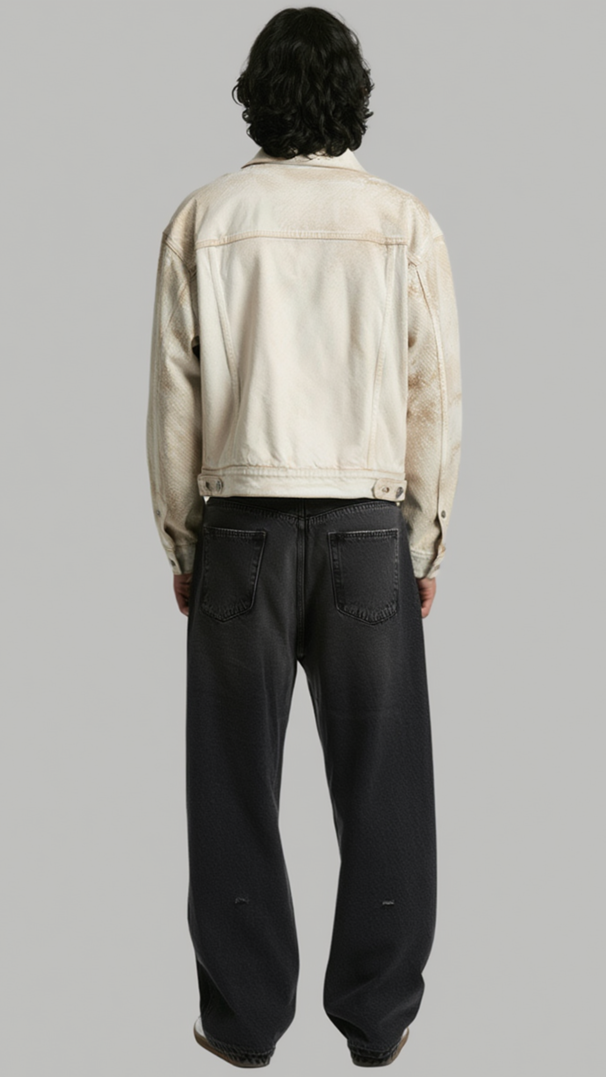 A model shown from the back wearing a cream-colored denim jacket and dark charcoal wide-leg jeans, set against a solid gray studio background.