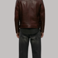 A man viewed from behind wearing a dark brown leather jacket, a black t-shirt, and black jeans, standing against a solid gray background.