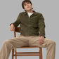 Person sitting on a chair wearing a green jacket and beige pants against a gray background
