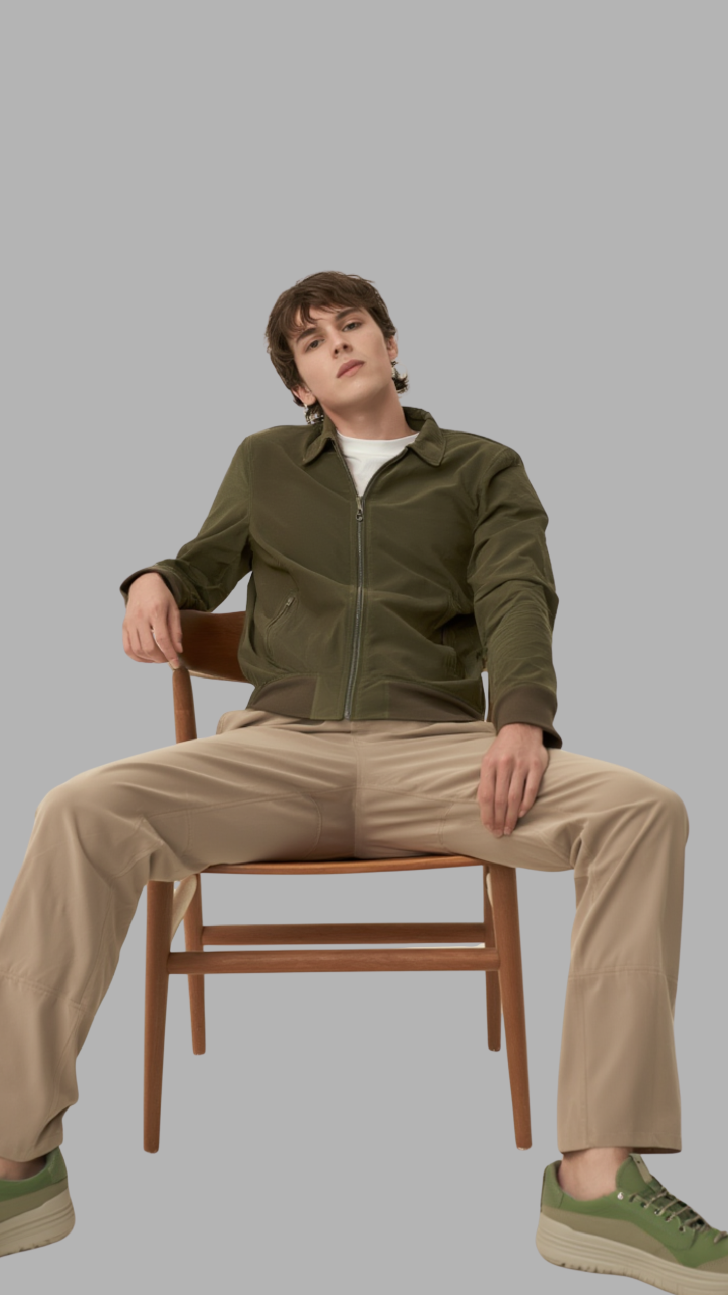 Person sitting on a chair wearing a green jacket and beige pants against a gray background