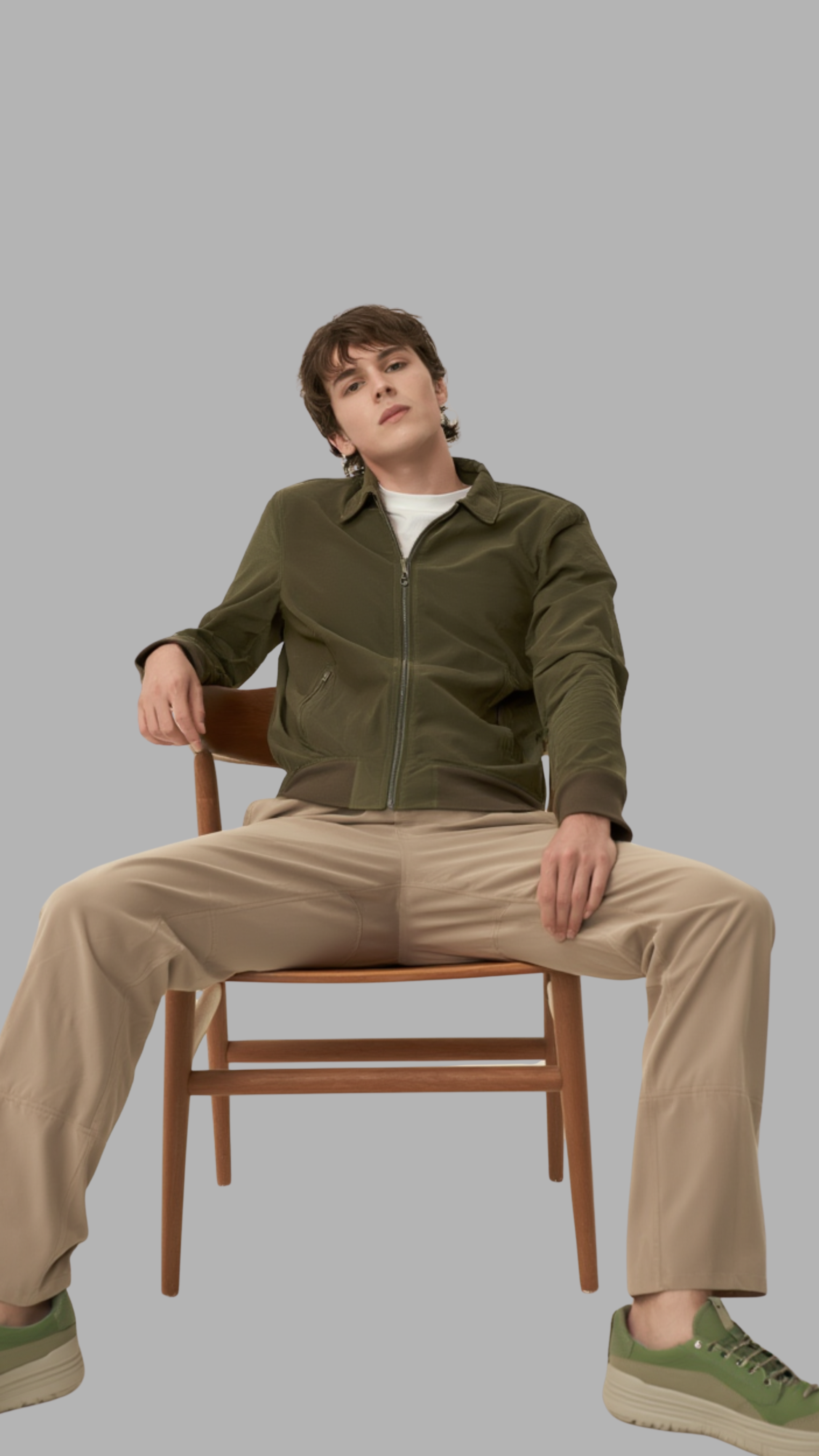 Person sitting on a chair wearing a green jacket and beige pants against a gray background