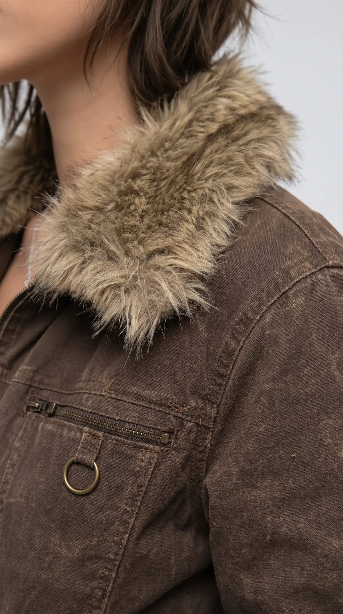 Close-up of a dark khaki distressed denim jacket showcasing its thick tan faux-fur collar, textured cotton denim, horizontal chest zipper and circular metal D-ring detail. The contrasting plush fibres, muted brown-green wash and visible rugged stitching bring depth to this cropped Y2K statement jacket, highlighting its vintage utility design and distinctive hardware for shoppers seeking alternative streetwear with character.