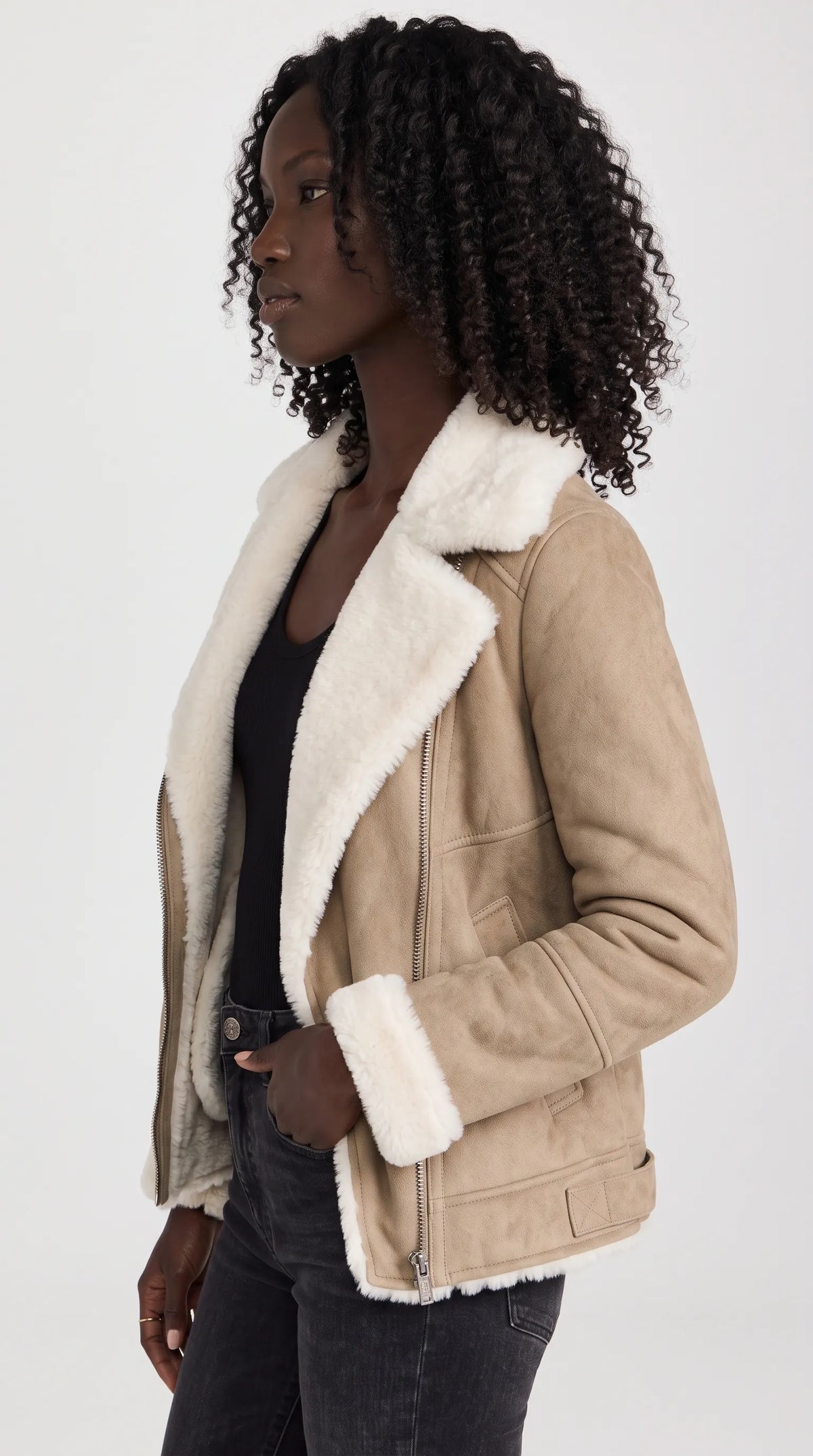 A taupe-beige suede shearling biker jacket brings a distinctive moto silhouette to everyday winter fashion, with a broad white plush collar, angled front zipper, streamlined side pockets and fitted cuffs. Styled with black denim, this statement outerwear piece balances rugged aviator character with clean, modern urban streetwear.
