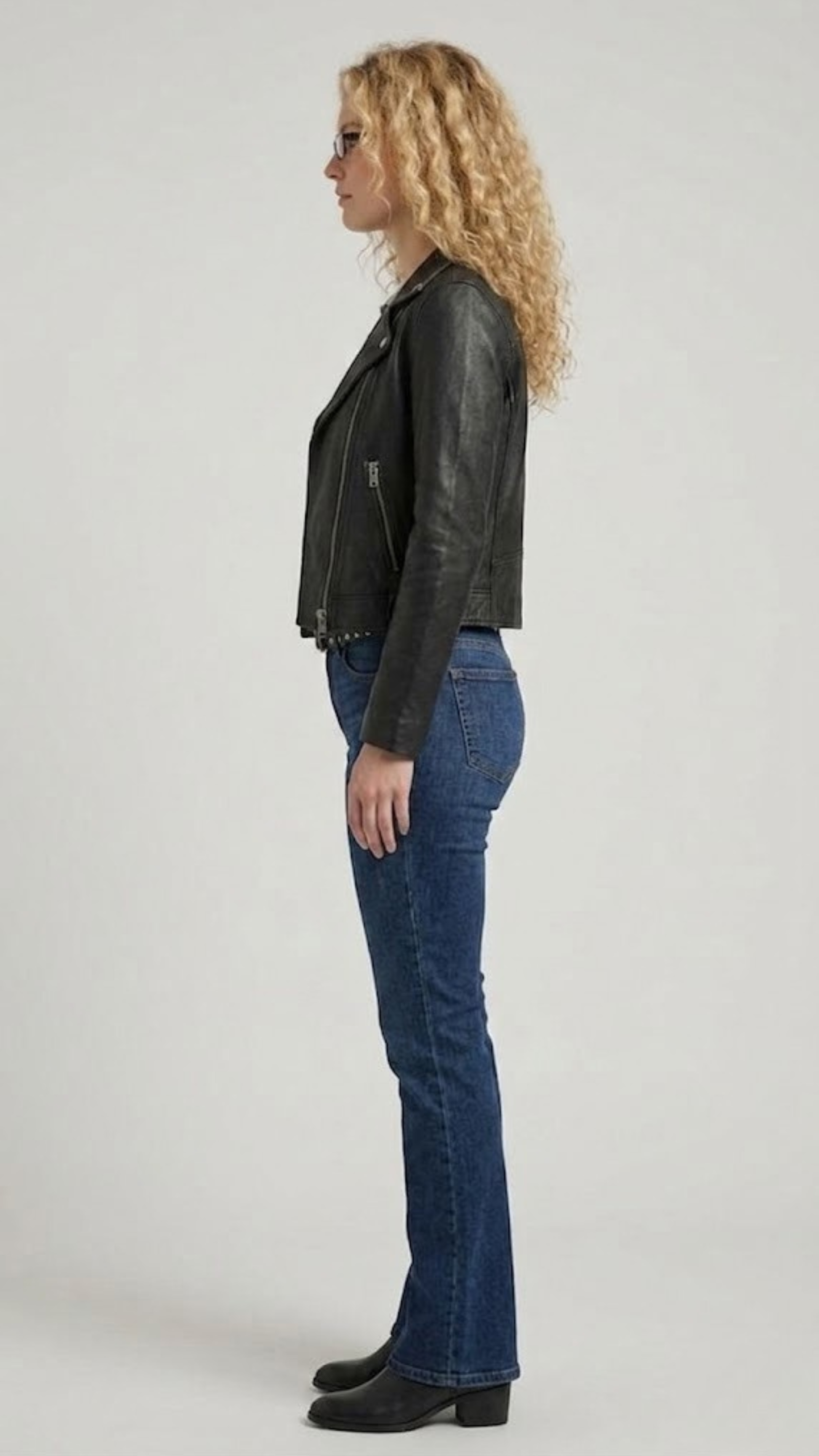 Full-length studio photography of a model wearing a stylish men's black leather motorcycle jacket paired with vintage jeans, showcasing a sleek streetwear look.