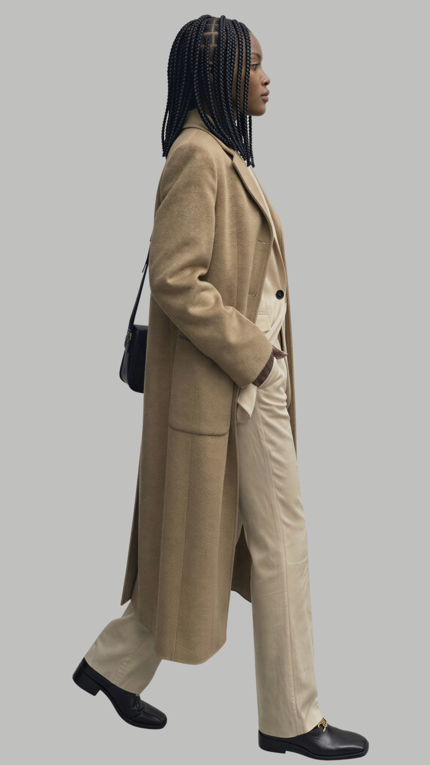 Woman wearing a beige coat and pants with a black handbag on a gray background