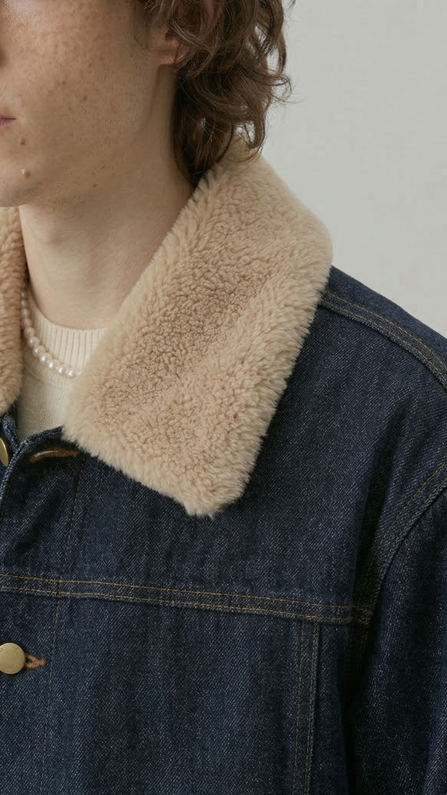 Close-up of a person wearing a dark indigo denim jacket with a thick beige sherpa fleece collar over a cream ribbed sweater.