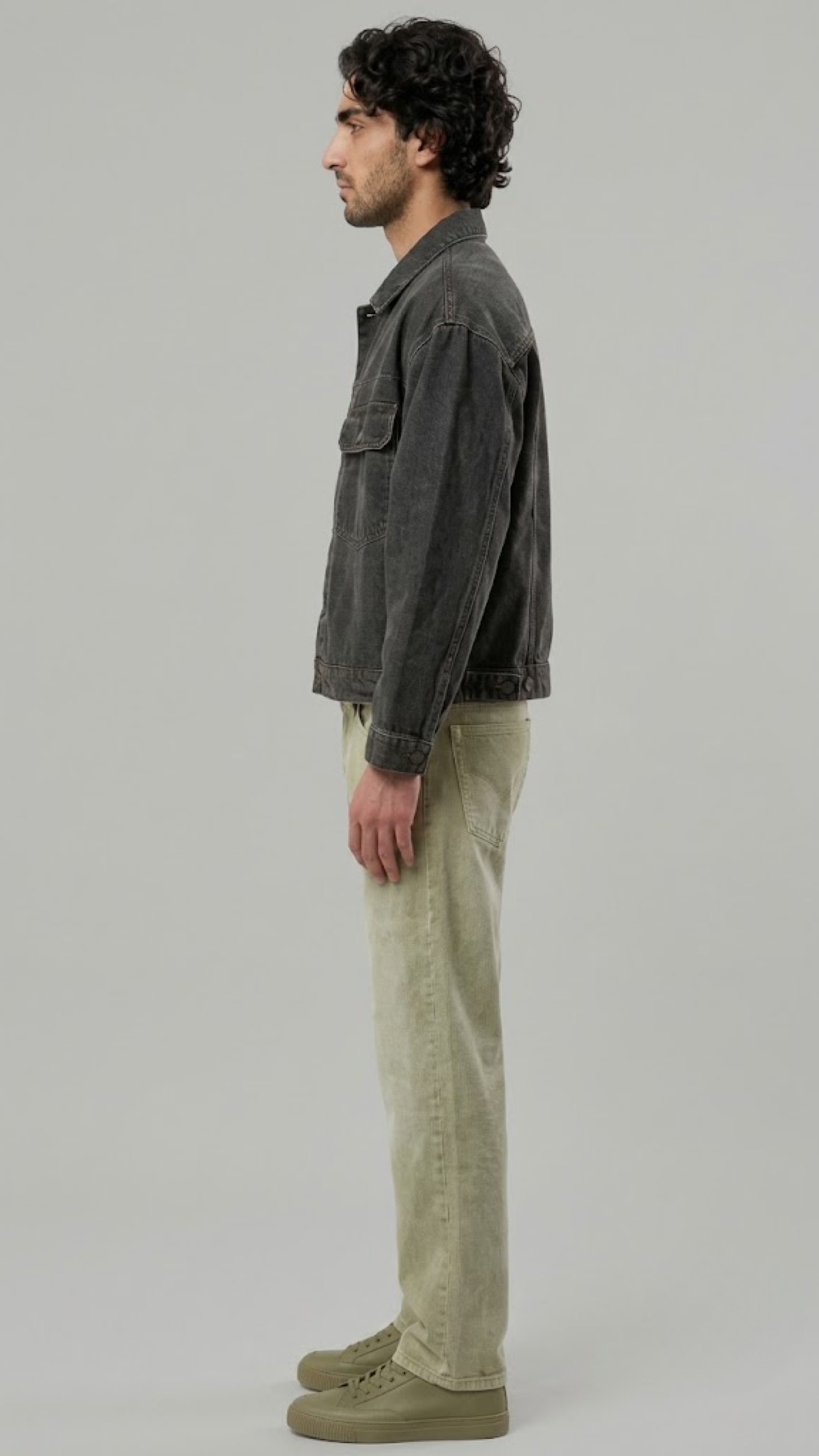 A man in profile wearing a black leather jacket with white stripes down the sleeves, blue jeans, and black leather boots, standing against a light gray studio background.