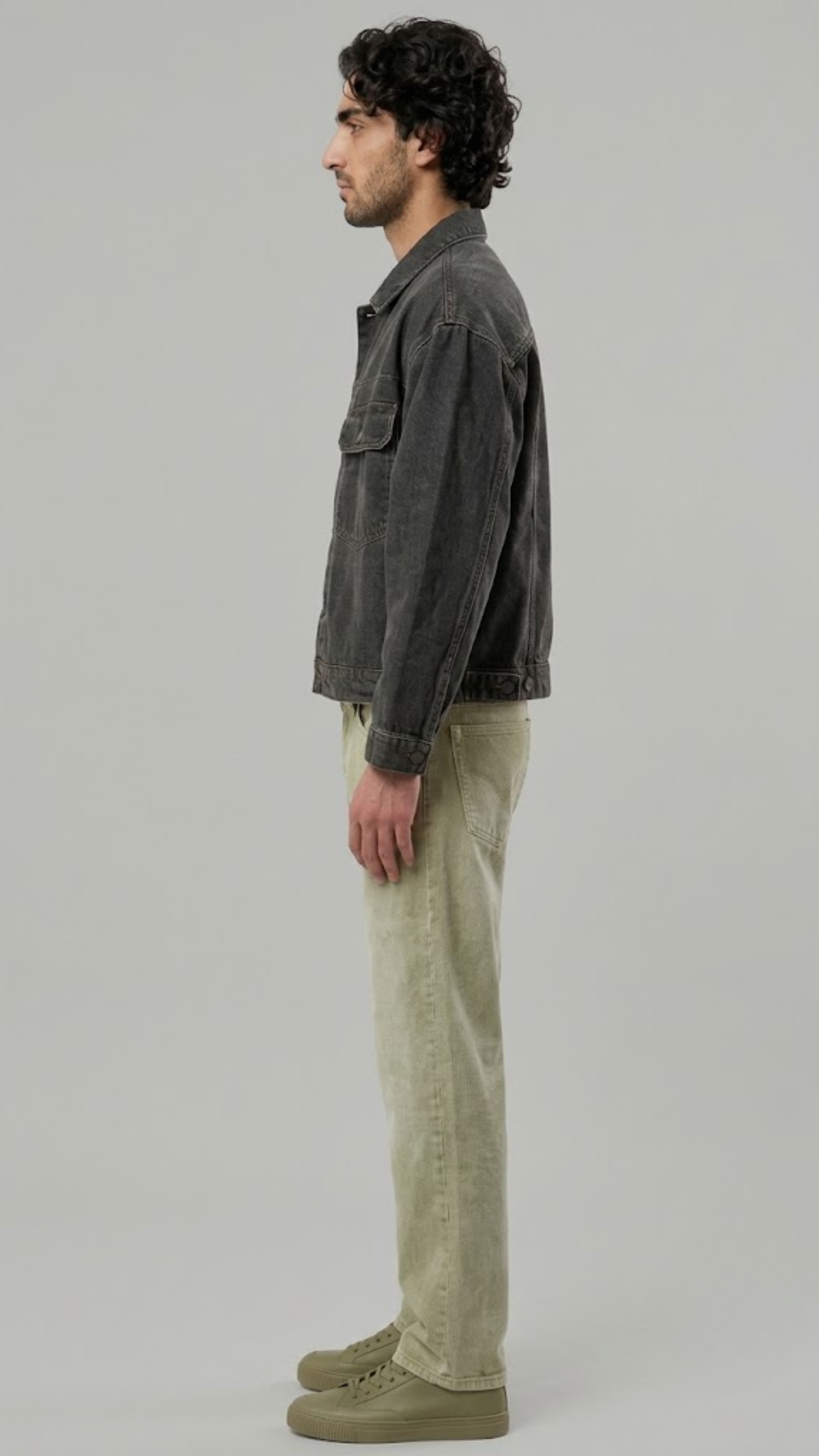 A man in profile wearing a black leather jacket with white stripes down the sleeves, blue jeans, and black leather boots, standing against a light gray studio background.