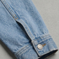 A close-up of a light blue denim jacket sleeve showing the buttoned cuff and raw hem, set against a neutral gray background.