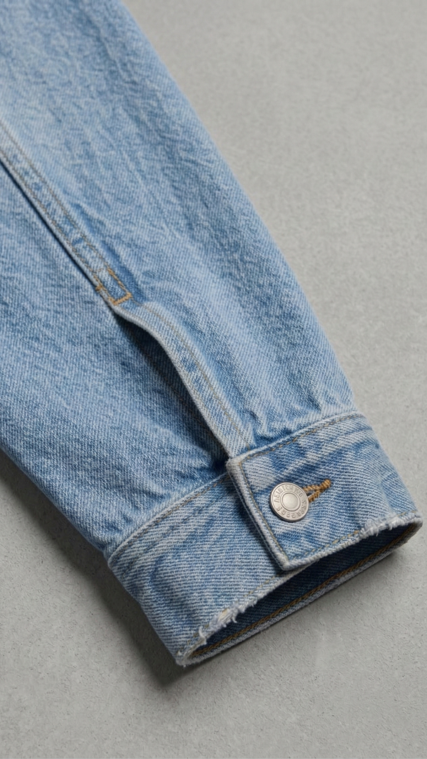 A close-up of a light blue denim jacket sleeve showing the buttoned cuff and raw hem, set against a neutral gray background.