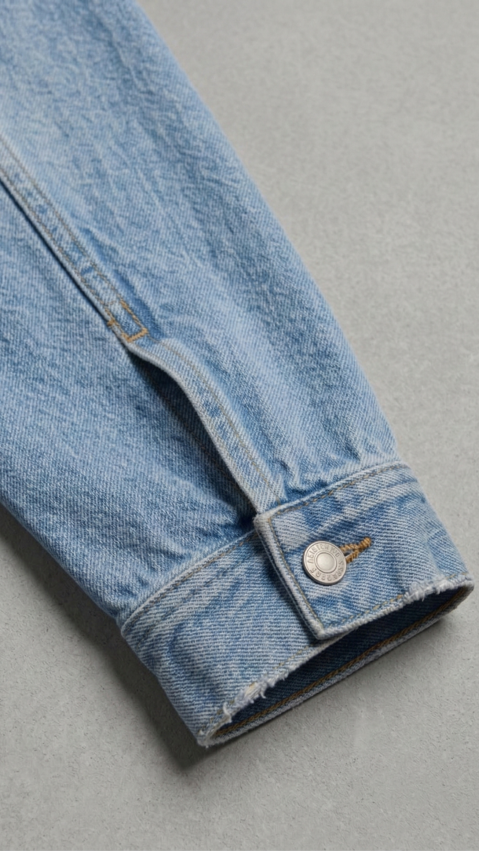A close-up of a light blue denim jacket sleeve showing the buttoned cuff and raw hem, set against a neutral gray background.