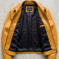 A mustard yellow leather jacket with a black satin lining, laid flat on a textured gray concrete surface. The brand label inside the collar reads 'JACKERO'.